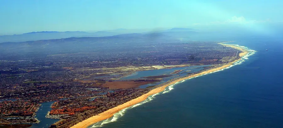 Aerial view looking down the coast over the Bolsa Chica wetlands, with the long sand beach and breaking surf on one side and neighborhoods on the other.